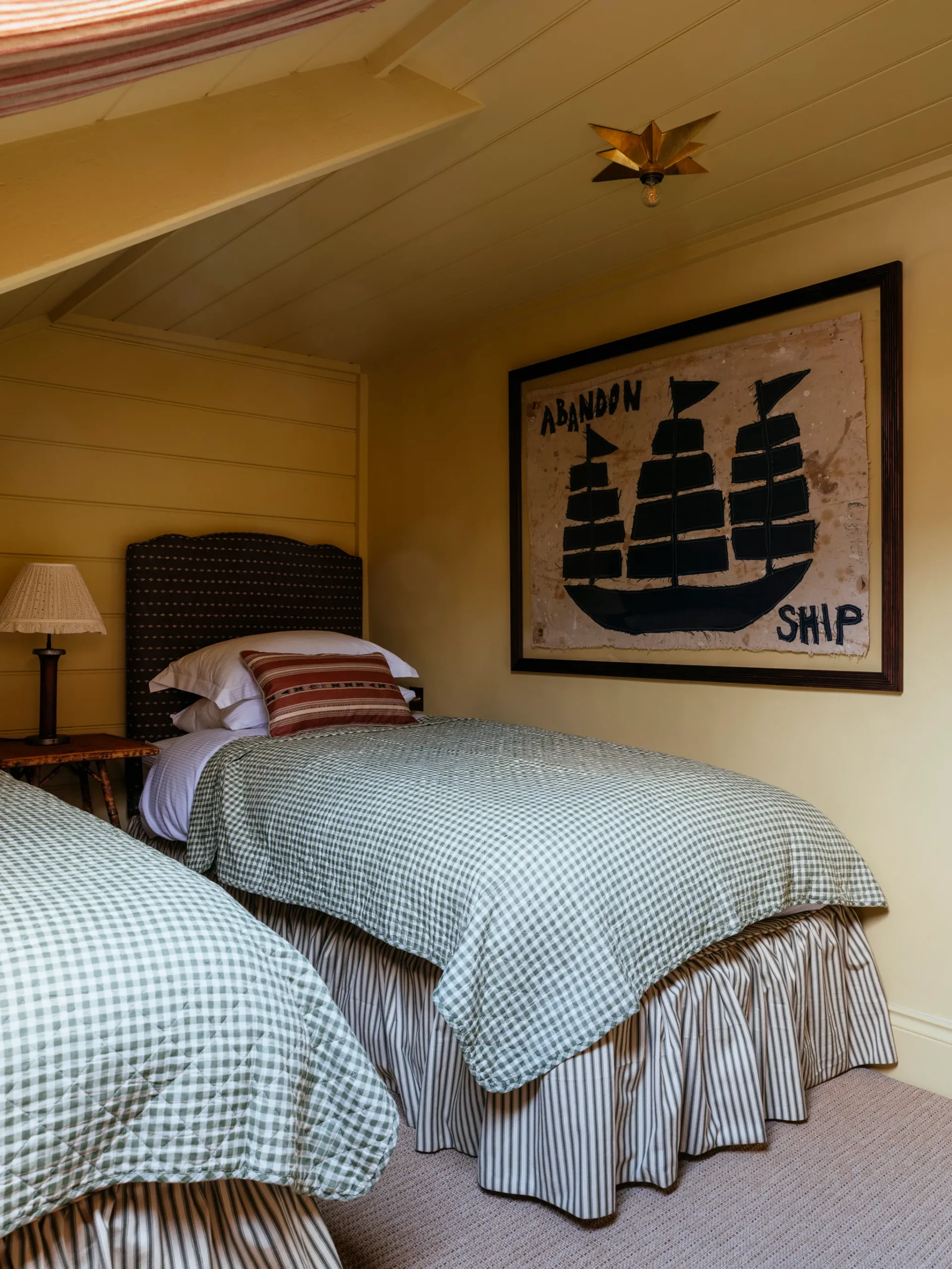 Small bedroom with wood plank walls painted yellow. Twin beds with gingham bedding. HÁM Interiors