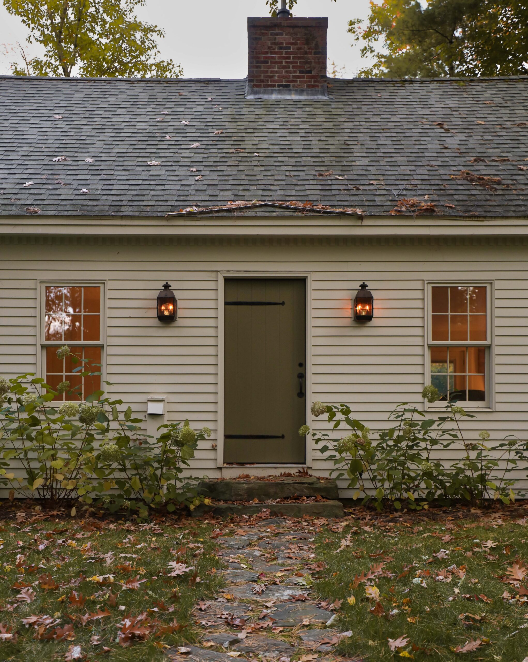 Cape Cod house with bronze lanterns and flickering flame light bulbs. Wood plank front door with iron strap hinges. Nadine Stay